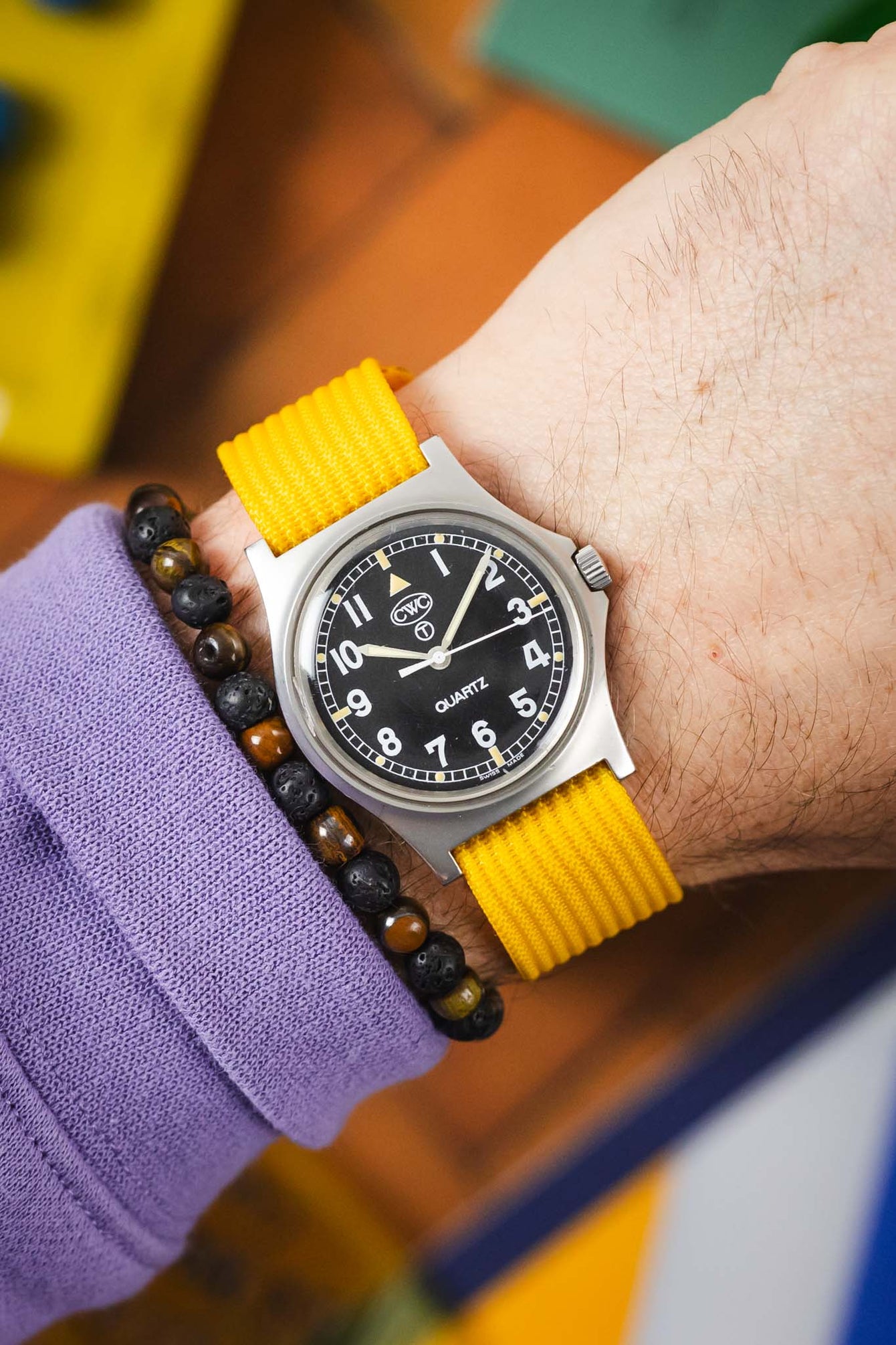 Close-up of a wrist wearing a silver analog watch with a black dial, white numerals, and a bright yellow ribbed nylon strap, paired with a dark beaded bracelet and a purple sleeve, set against a colorful blurred background.