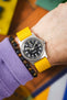 Close-up of a wrist wearing a silver analog watch with a black dial, white numerals, and a bright yellow ribbed nylon strap, paired with a dark beaded bracelet and a purple sleeve, set against a colorful blurred background.