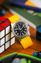 Close-up of a wrist wearing a silver watch with a black face and bold white numbers on a bright yellow fabric strap, paired with a beaded bracelet. The background is blurred with warm tones of orange, yellow, and green.
