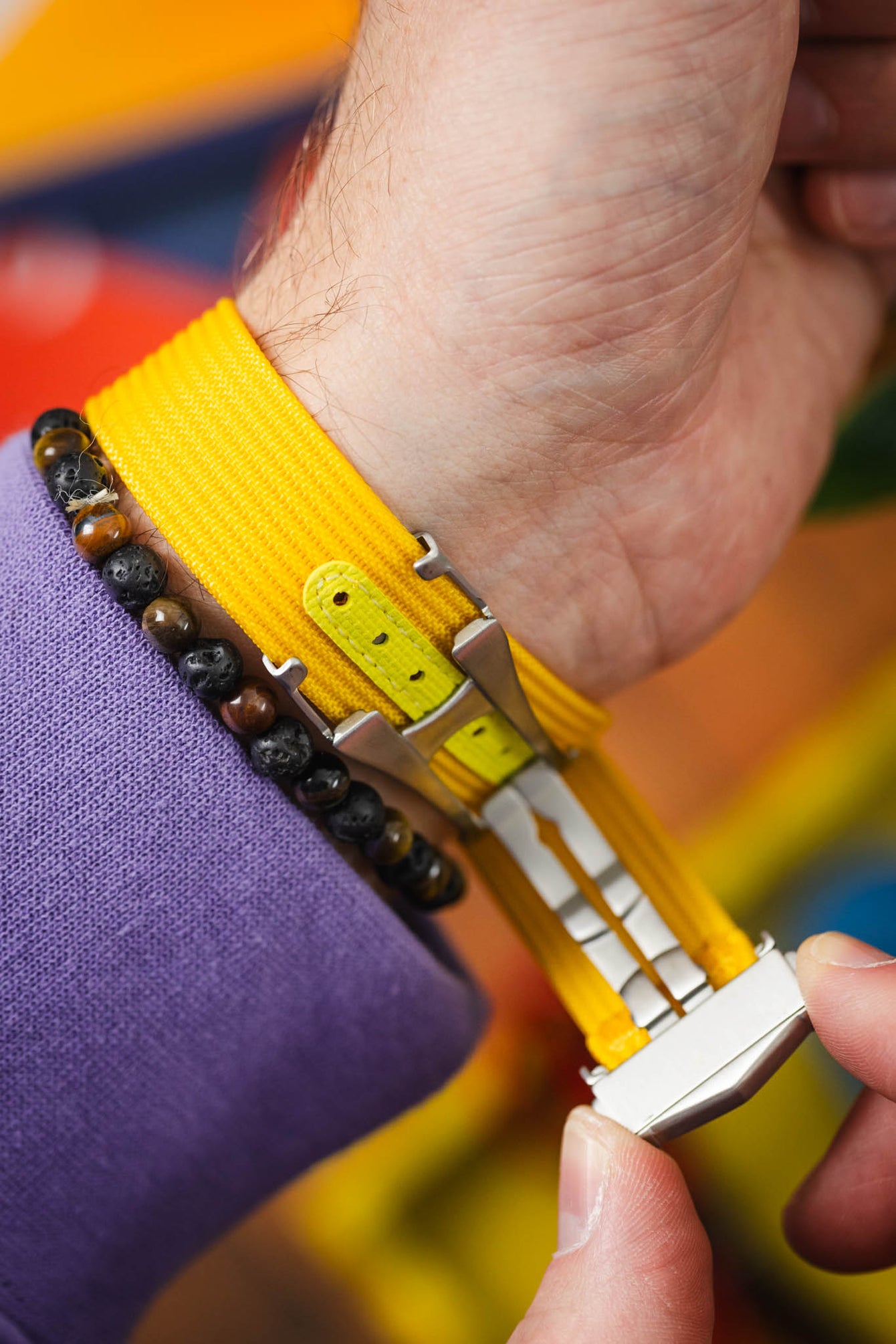 Close-up of a wrist showing the underside of a bright yellow ribbed fabric watch strap with a metal clasp being fastened by hand, alongside a dark beaded bracelet and a purple sleeve, against a colorful blurred background.