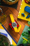 Flat lay of a silver analog wristwatch with a black dial and bright yellow fabric strap placed on a red-and-black cube, surrounded by colorful everyday items including blue wireless earbuds, a yellow tray, a green notebook with a leaf design, a keychain, and a plant, all on a wooden surface.