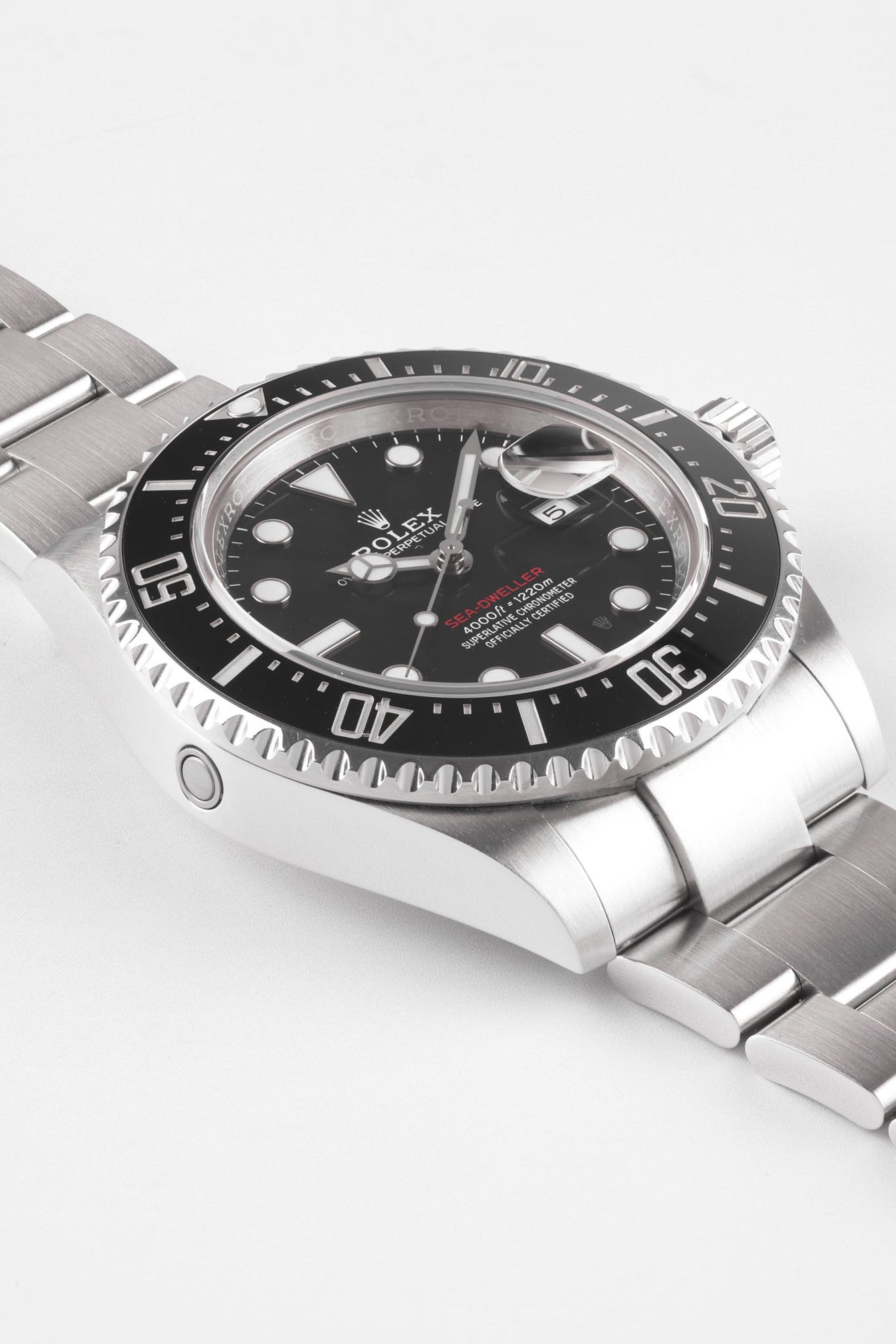 Angled close-up of a stainless steel Rolex Sea-Dweller watch showing the black dial, date magnifier, rotating bezel, and brushed metal bracelet against a plain background.