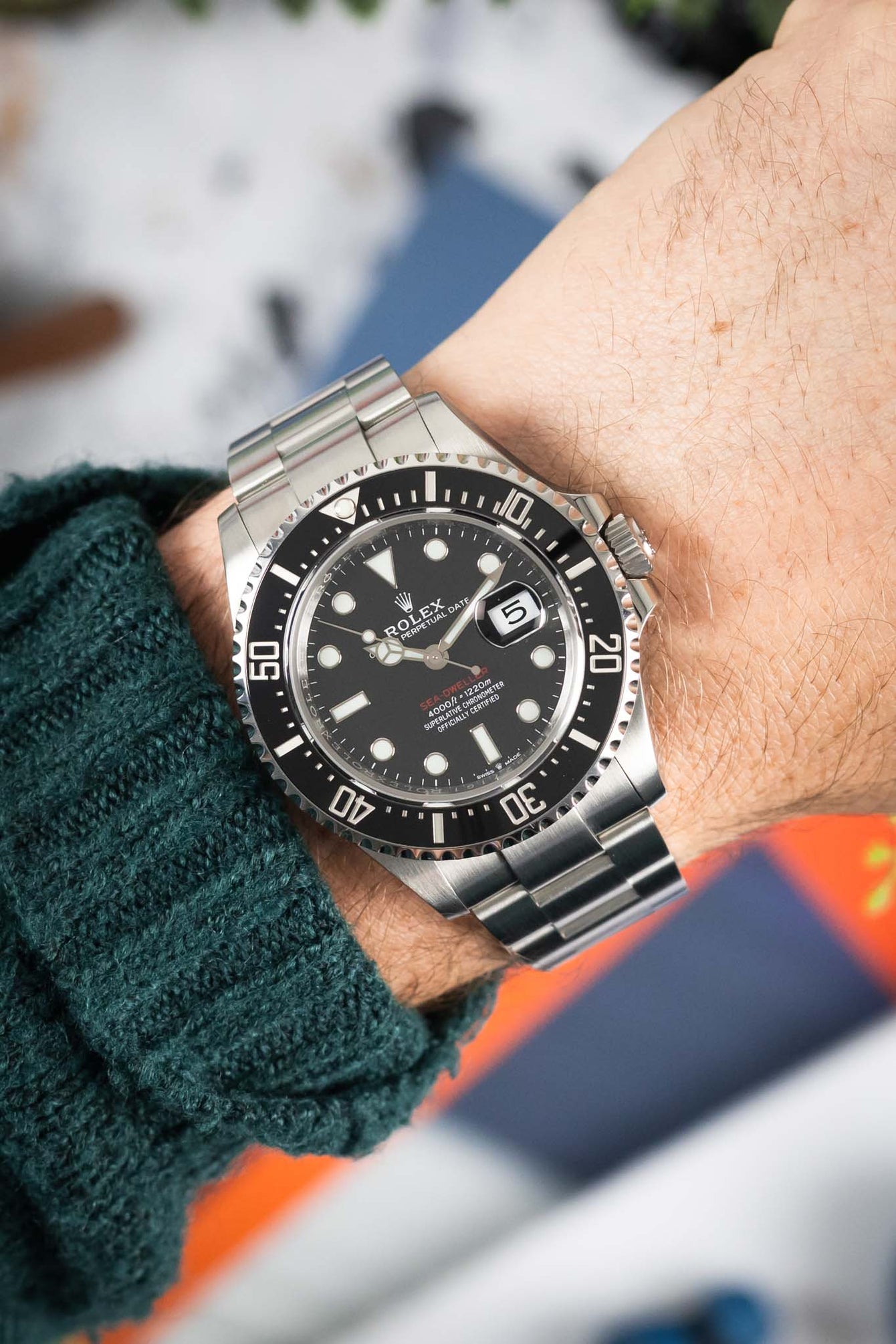 Stainless steel Rolex Sea-Dweller dive watch with black dial and date display worn on a wrist, shown in close-up with a green knit sleeve and blurred background.