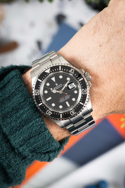 Stainless steel Rolex Sea-Dweller dive watch with black dial and date display worn on a wrist, shown in close-up with a green knit sleeve and blurred background.