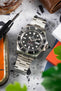 Stainless steel Rolex Sea-Dweller watch with black dial and date display, placed among workshop tools and objects on a speckled surface.