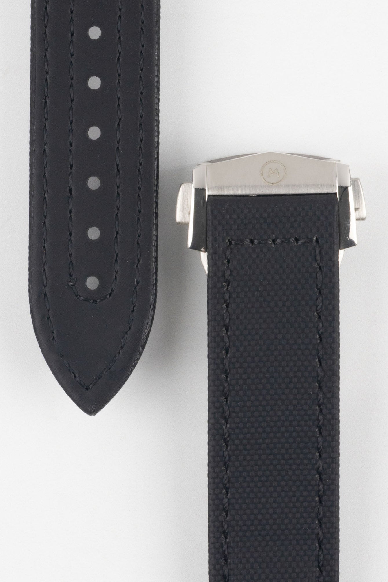 Close-up of a black textured watch strap showing two sections: one with evenly spaced adjustment holes and stitched edges, and the other attached to a polished silver deployant clasp with a subtle engraved logo, set against a light grey background.