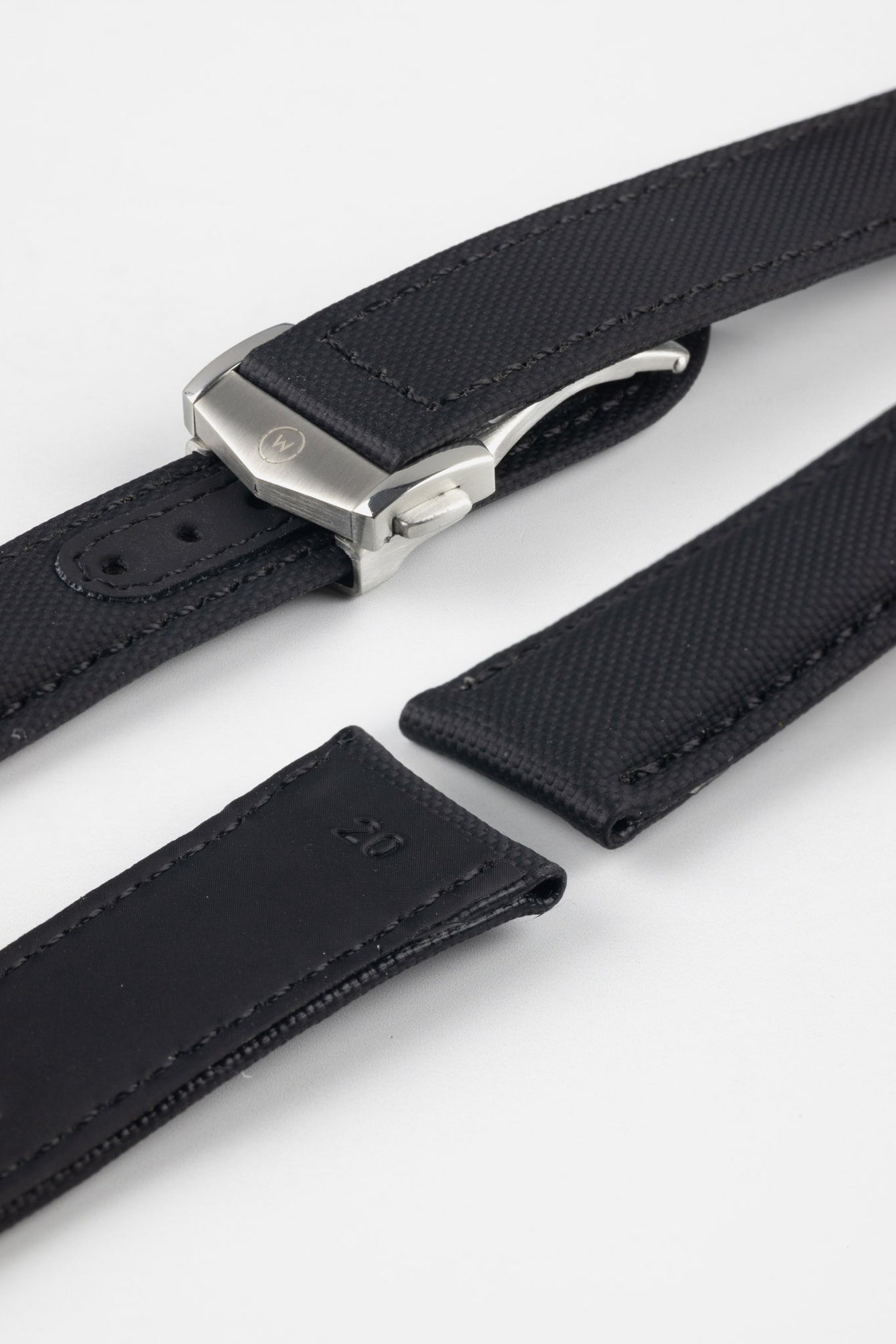 Angled close-up of a black textured watch strap featuring a brushed silver deployant clasp with an engraved logo, alongside separate strap sections with visible stitching and adjustment holes, displayed on a light grey background.