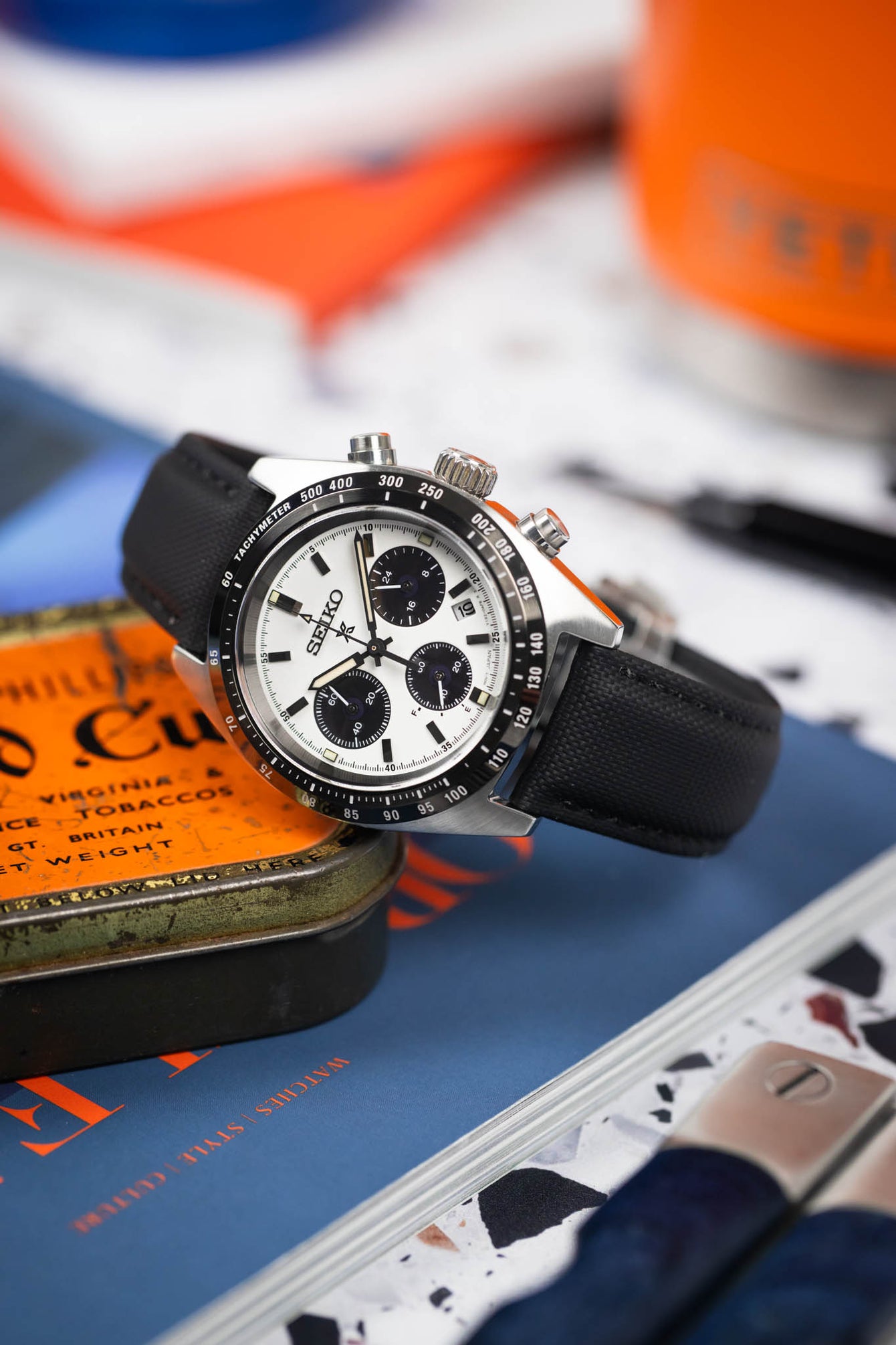 Close-up of a Seiko chronograph with a white dial and black subdials on a black strap, resting on a tin and magazines.