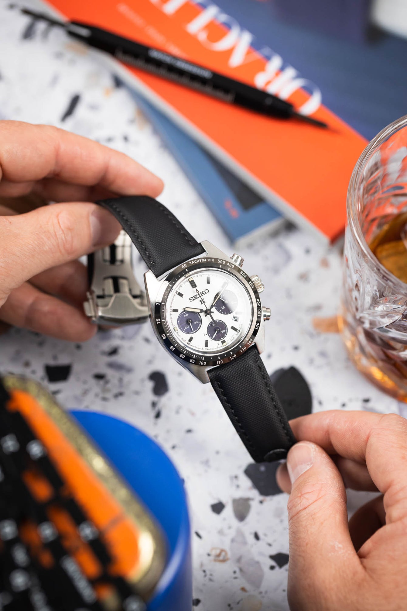 Close-up of a Seiko chronograph with a white dial on a black strap, held in hands above a desk with books and a glass.