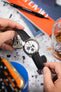 Close-up of a Seiko chronograph with a white dial on a black strap, held in hands above a desk with books and a glass.