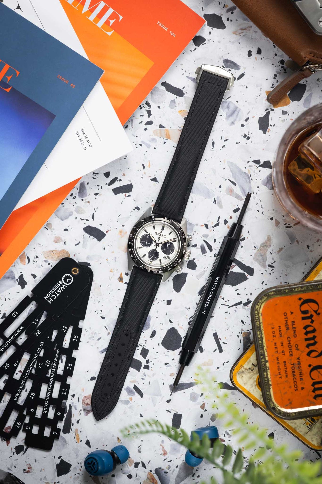 Flat lay of a Seiko chronograph on a black textured strap, surrounded by tools, magazines, a drink, and accessories on a speckled surface.