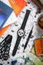 Flat lay of a Seiko chronograph on a black textured strap, surrounded by tools, magazines, a drink, and accessories on a speckled surface.