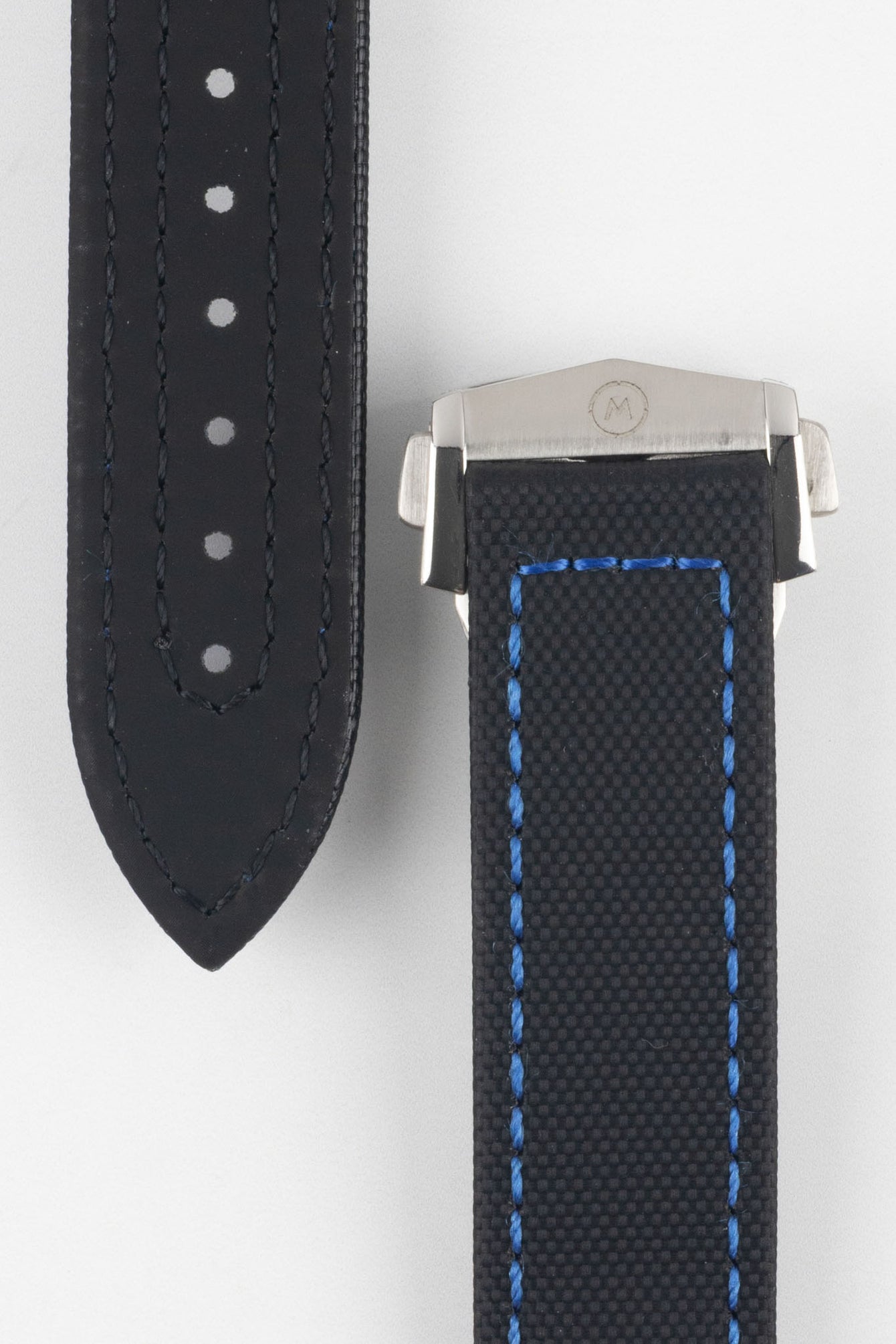 Close-up of a black textured watch strap with blue contrast stitching, showing one section with evenly spaced adjustment holes and the other attached to a brushed silver deployant clasp with an engraved logo, set against a light grey background.