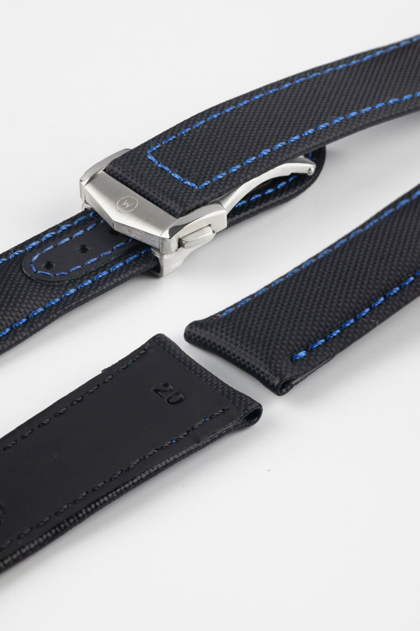 Angled close-up of a black textured watch strap with blue contrast stitching, featuring a brushed silver deployant clasp with an engraved logo, alongside separate strap sections, all displayed on a light grey background.