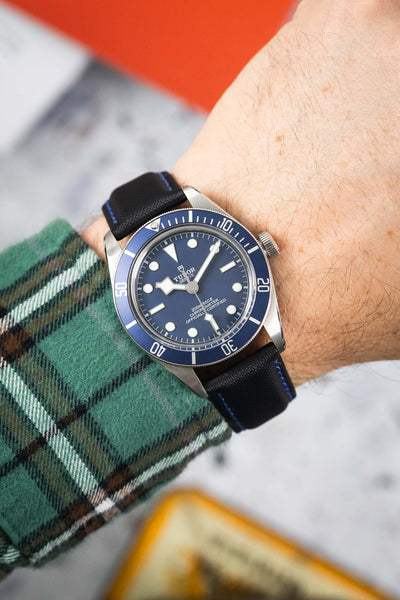 Close-up of a Tudor dive watch with a blue dial and rotating bezel, worn on a wrist with a black textured strap featuring blue stitching. The watch shows luminous hour markers and “200m:660ft CHRONOMETER OFFICIALLY CERTIFIED” text, while a green plaid sleeve and softly blurred background frame the scene.