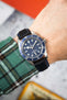 Close-up of a Tudor dive watch with a blue dial and rotating bezel, worn on a wrist with a black textured strap featuring blue stitching. The watch shows luminous hour markers and “200m:660ft CHRONOMETER OFFICIALLY CERTIFIED” text, while a green plaid sleeve and softly blurred background frame the scene.