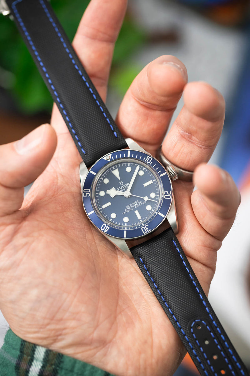 Close-up of a Tudor dive watch with a blue dial and bezel, held in a hand and fitted to a black textured strap with blue stitching. The luminous markers and “200m:660ft CHRONOMETER OFFICIALLY CERTIFIED” text are visible, while the softly blurred background adds contrast to the watch’s crisp details.