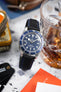 Close-up of a Tudor dive watch with a blue dial and rotating bezel on a black textured strap with blue stitching, resting beside a glass of amber drink. The watch’s luminous markers and chronometer text are visible, while the softly blurred background includes a vintage tin and colorful printed materials.