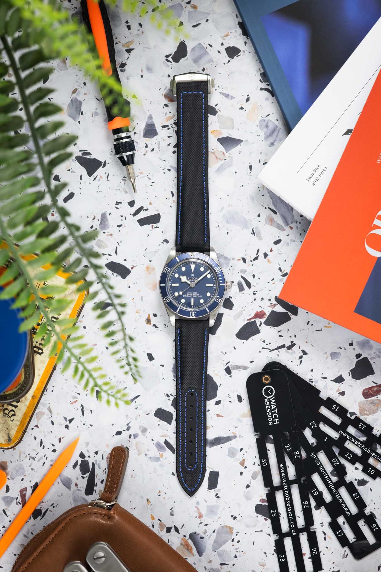 Flat lay of a Tudor dive watch with a blue dial and bezel on a black textured strap with blue stitching, arranged on a speckled surface. The scene includes watch tools, a leather pouch, measuring gauges, magazines, and greenery, creating a workshop-style composition with contrasting blue, orange, and neutral tones.