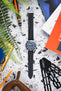 Flat lay of a Tudor dive watch with a blue dial and bezel on a black textured strap with blue stitching, arranged on a speckled surface. The scene includes watch tools, a leather pouch, measuring gauges, magazines, and greenery, creating a workshop-style composition with contrasting blue, orange, and neutral tones.