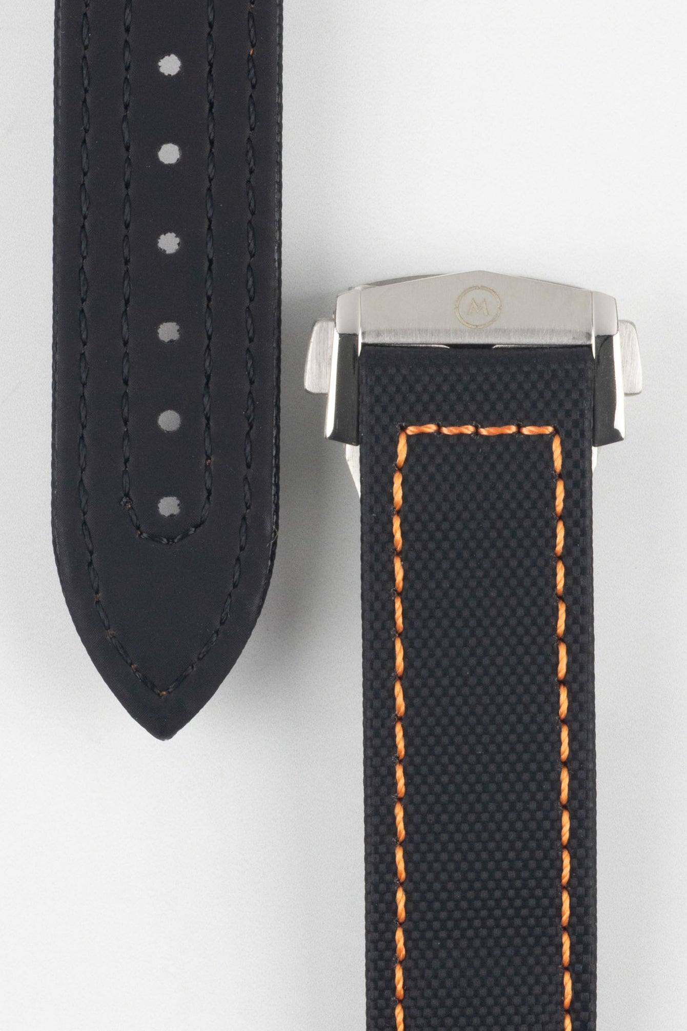 Close-up of a black textured watch strap with orange contrast stitching, showing one section with evenly spaced adjustment holes and the other attached to a brushed silver deployant clasp with an engraved logo, set against a light grey background.