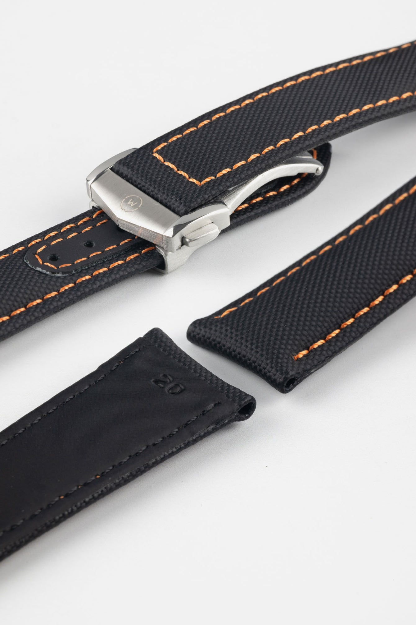 Angled close-up of a black textured watch strap with orange contrast stitching, featuring a brushed silver deployant clasp with an engraved logo, alongside separate strap sections, all displayed on a light grey background.