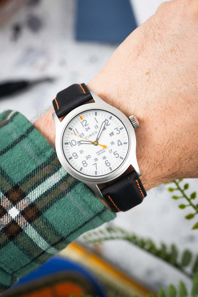 Close-up of a Timex field watch with a white dial and orange seconds hand, worn on a wrist with a black textured strap featuring orange stitching. The dial shows full Arabic numerals and 24-hour markings, while a green plaid sleeve and blurred workshop-style background frame the watch.