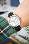 Close-up of a Timex field watch with a white dial and orange seconds hand, worn on a wrist with a black textured strap featuring orange stitching. The dial shows full Arabic numerals and 24-hour markings, while a green plaid sleeve and blurred workshop-style background frame the watch.