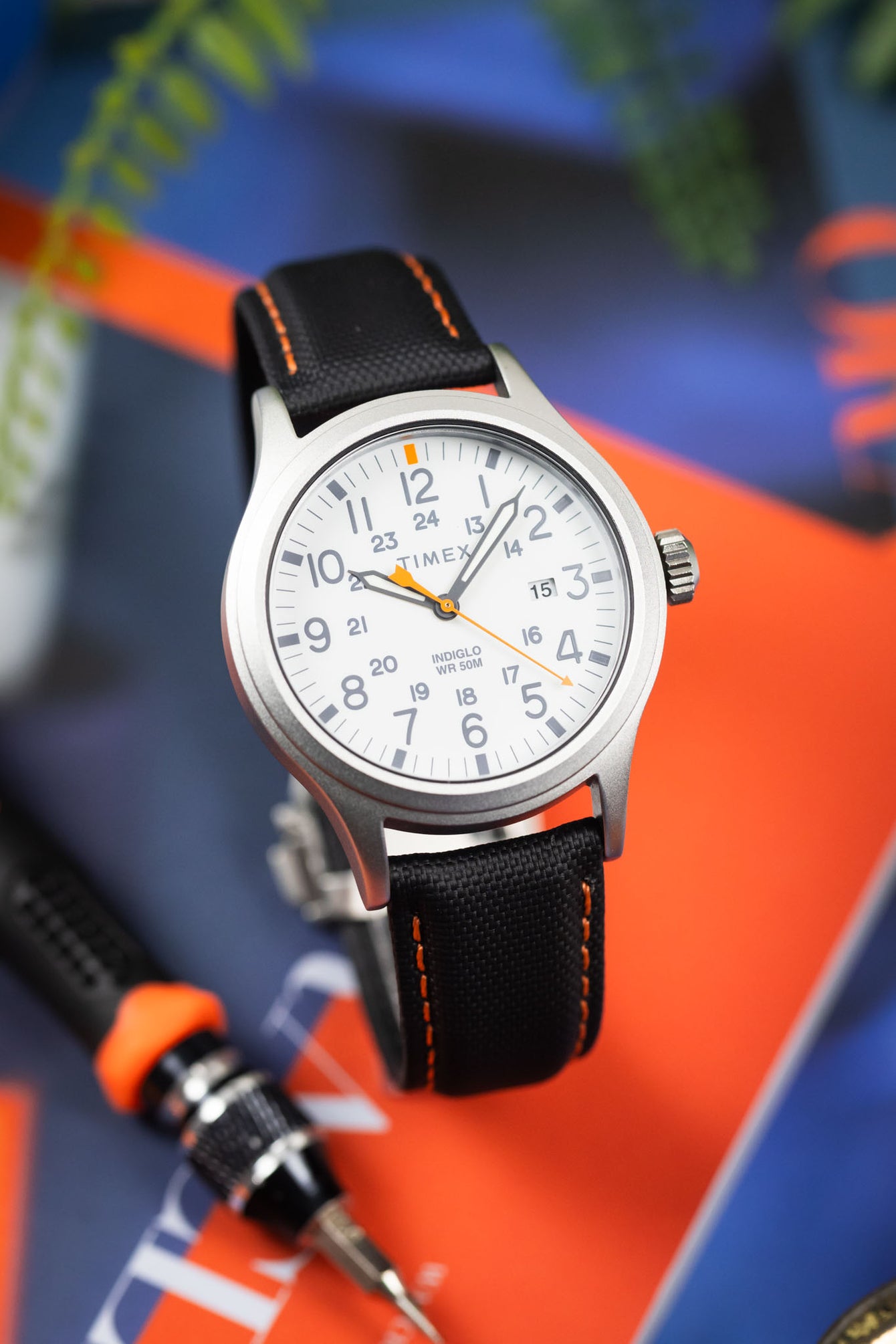 Close-up of a Timex field watch with a white dial and orange accents on a black textured strap with orange stitching, angled against a blurred background of bold orange and blue surfaces. The dial features full Arabic numerals, 24-hour markings, and “INDIGLO WR 50M” text, while the soft lighting emphasizes the brushed case and woven strap texture.