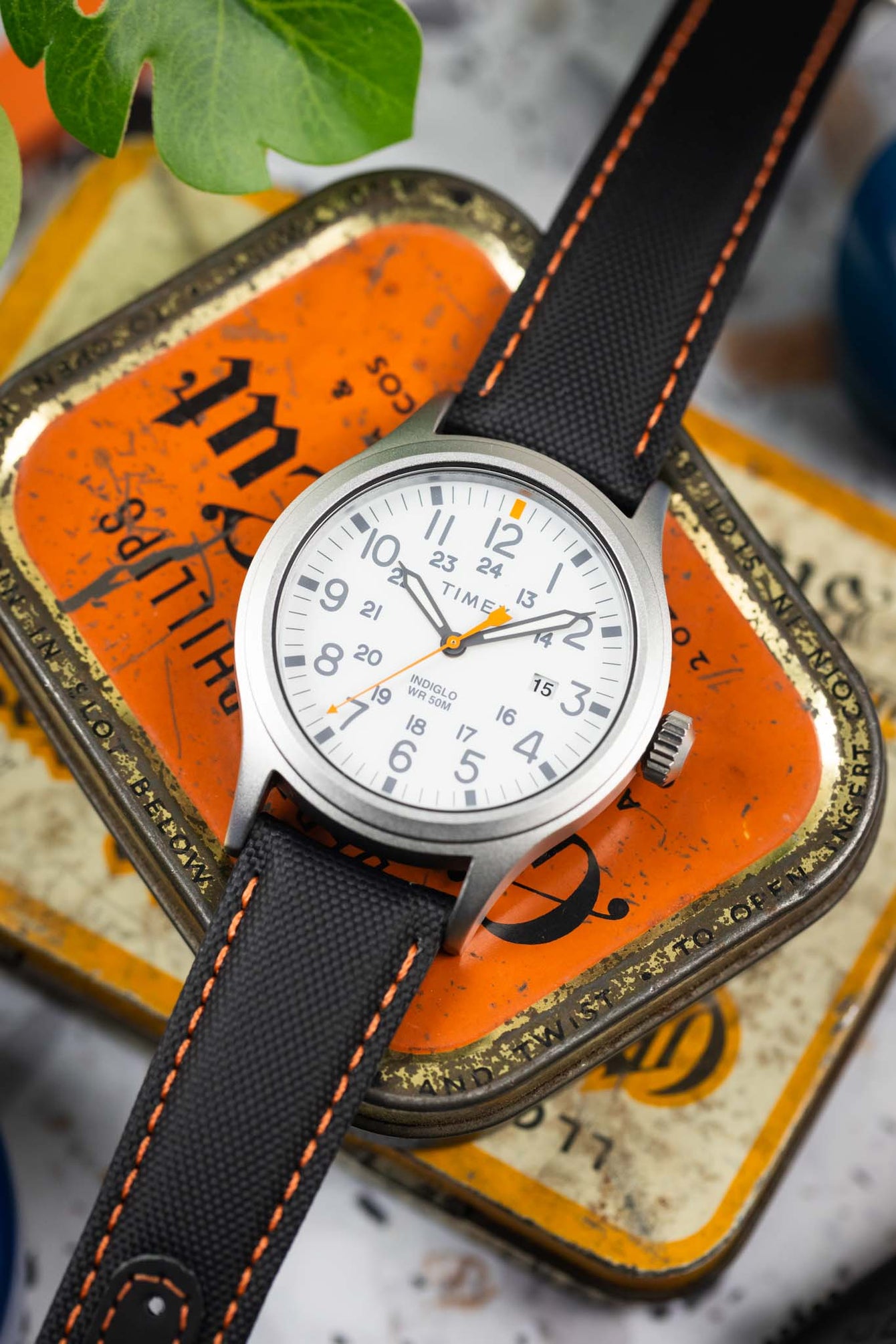 Close-up of a Timex field watch with a white dial and orange accents on a black textured strap with orange stitching, resting on a vintage-style orange tin. The dial features full Arabic numerals, 24-hour markings, a date window, and “INDIGLO WR 50M” text, while a green leaf and softly blurred background add contrast and depth.
