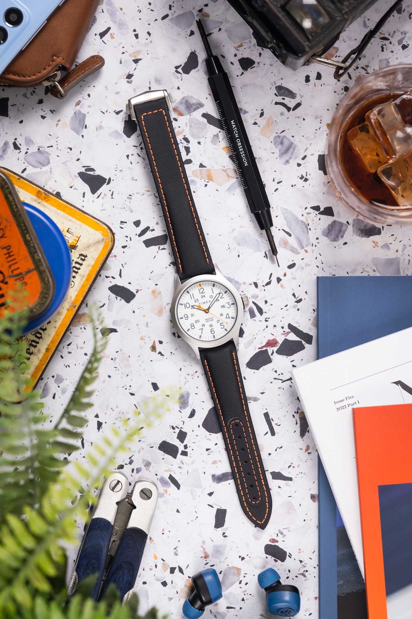 Flat lay of a Timex field watch with a white dial and orange accents on a black textured strap with orange stitching, arranged on a speckled surface. The scene is styled with watch tools, a glass with iced drink, a leather pouch, earbuds, magazines, and greenery, creating a casual workshop-inspired setting with a mix of warm and cool tones.