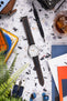 Flat lay of a Timex field watch with a white dial and orange accents on a black textured strap with orange stitching, arranged on a speckled surface. The scene is styled with watch tools, a glass with iced drink, a leather pouch, earbuds, magazines, and greenery, creating a casual workshop-inspired setting with a mix of warm and cool tones.