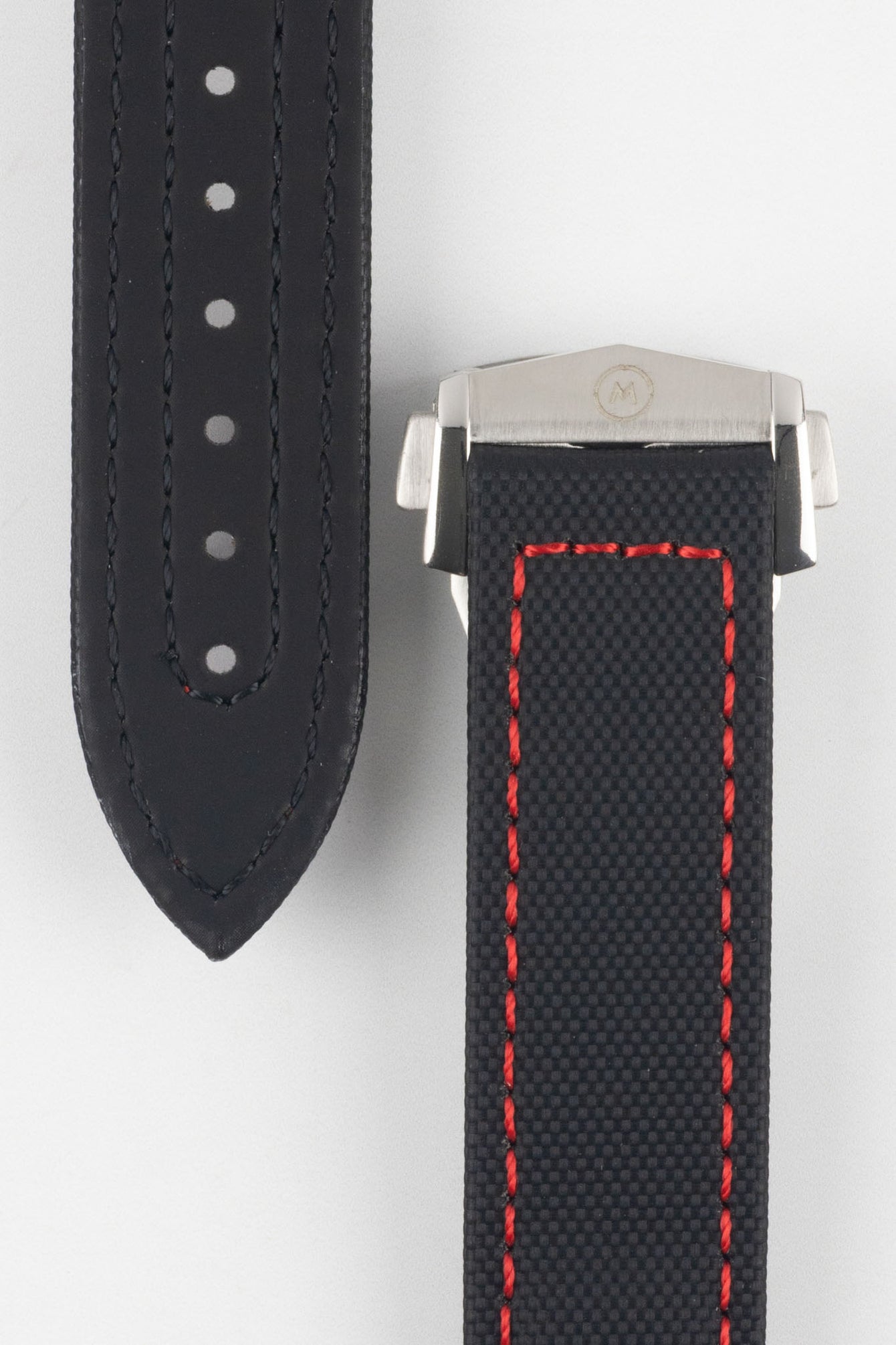 Close-up of a black textured watch strap with red contrast stitching, showing one section with evenly spaced adjustment holes and the other attached to a brushed silver deployant clasp with an engraved logo, set against a light grey background.