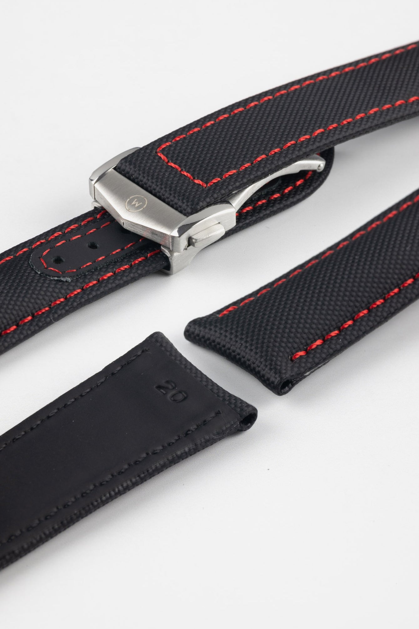 Angled close-up of a black textured watch strap with red contrast stitching, featuring a brushed silver deployant clasp with an engraved logo, alongside separate strap sections, all displayed on a light grey background.