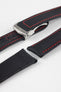 Angled close-up of a black textured watch strap with red contrast stitching, featuring a brushed silver deployant clasp with an engraved logo, alongside separate strap sections, all displayed on a light grey background.