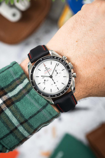 Close-up of an Omega Speedmaster Professional with a white dial and black bezel, worn on a textured black strap with red stitching. The chronograph subdials and tachymeter scale are clearly visible, while a green plaid sleeve frames the wrist against a softly blurred background with warm-toned objects.