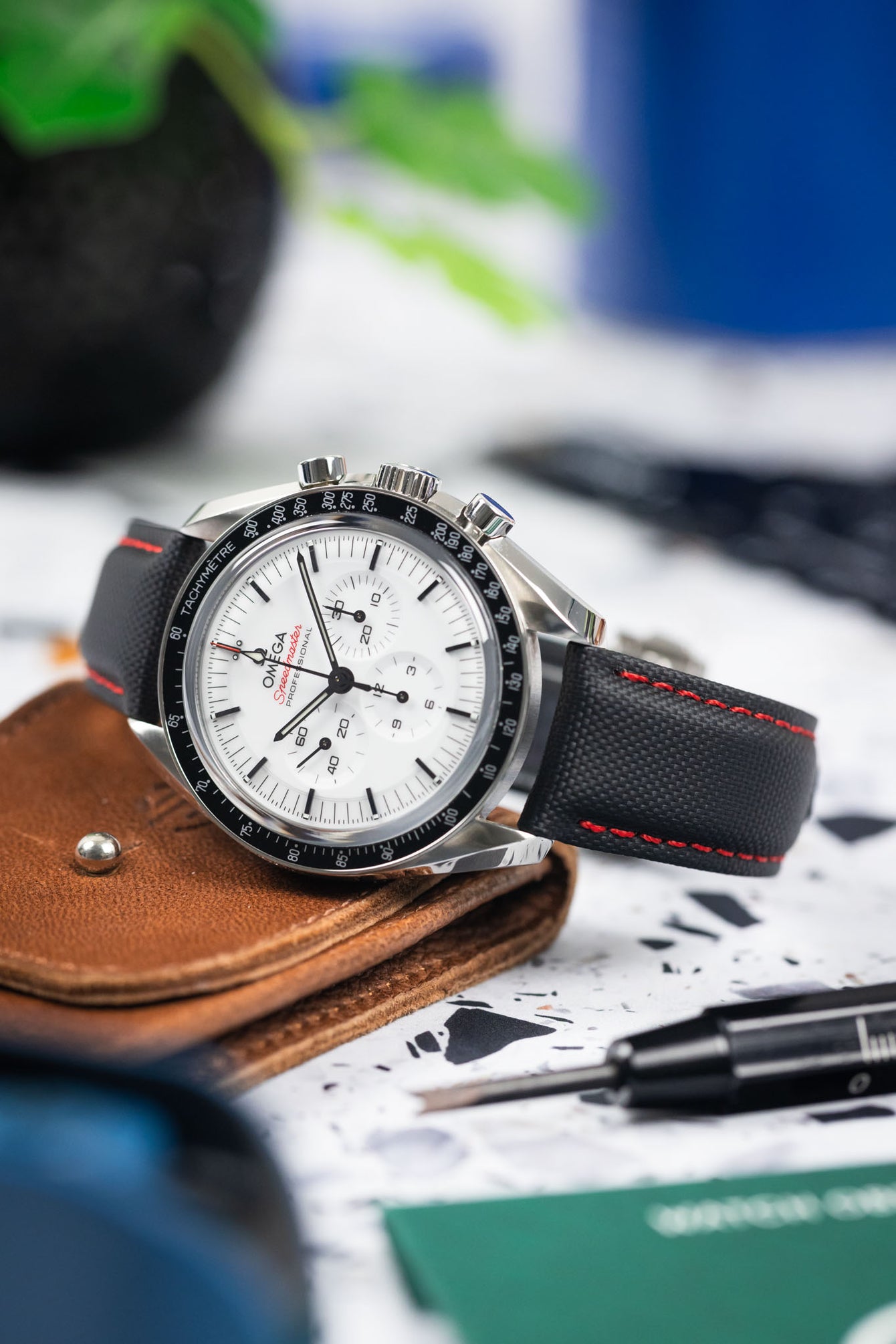 Close-up of an Omega Speedmaster Professional with a white dial and black bezel resting on a brown leather case. The watch is paired with a black textured strap featuring red stitching, while a small tool and blurred everyday objects in the background create a workshop-style setting.