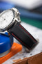 Macro close-up of a black textured watch strap with red contrast stitching attached to a polished steel watch case. The shallow depth of field highlights the woven sailcloth-like material and stitching detail, while the blurred background shows hints of blue, green, and a brown leather surface.