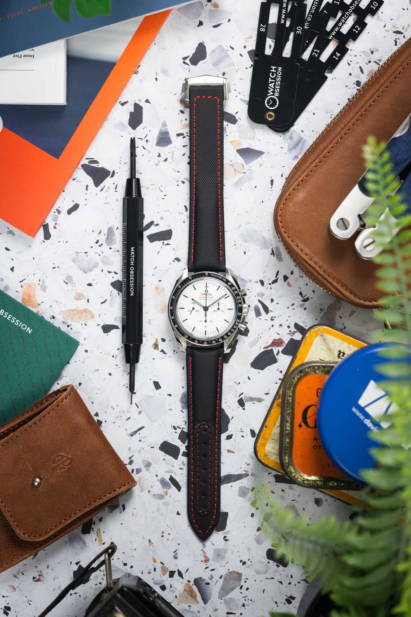 Flat lay of an Omega Speedmaster watch with a white dial on a black textured strap featuring red stitching, placed on a speckled surface. The scene is styled with watch tools, a leather pouch, a tin, magazines, and greenery, creating a workshop-inspired composition with a mix of warm and cool tones.