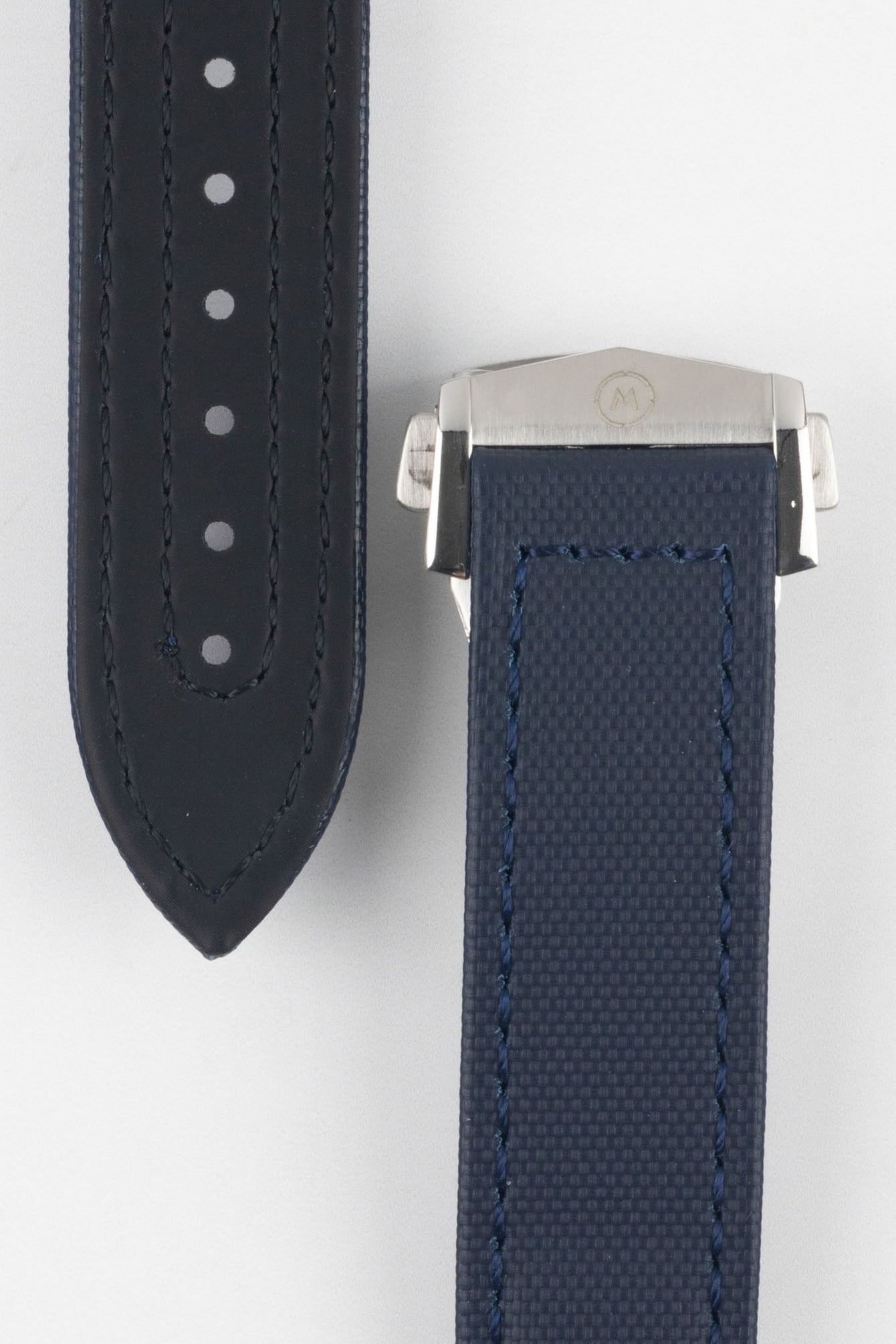 Close-up of a dark blue textured watch strap with matching blue stitching, showing one section with a row of evenly spaced adjustment holes and the other attached to a brushed silver deployant clasp with an engraved logo, set against a light grey background.