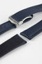 Angled close-up of a dark blue textured watch strap with matching blue stitching, featuring a brushed silver deployant clasp with an engraved logo, alongside separate strap pieces, all arranged on a light grey background.