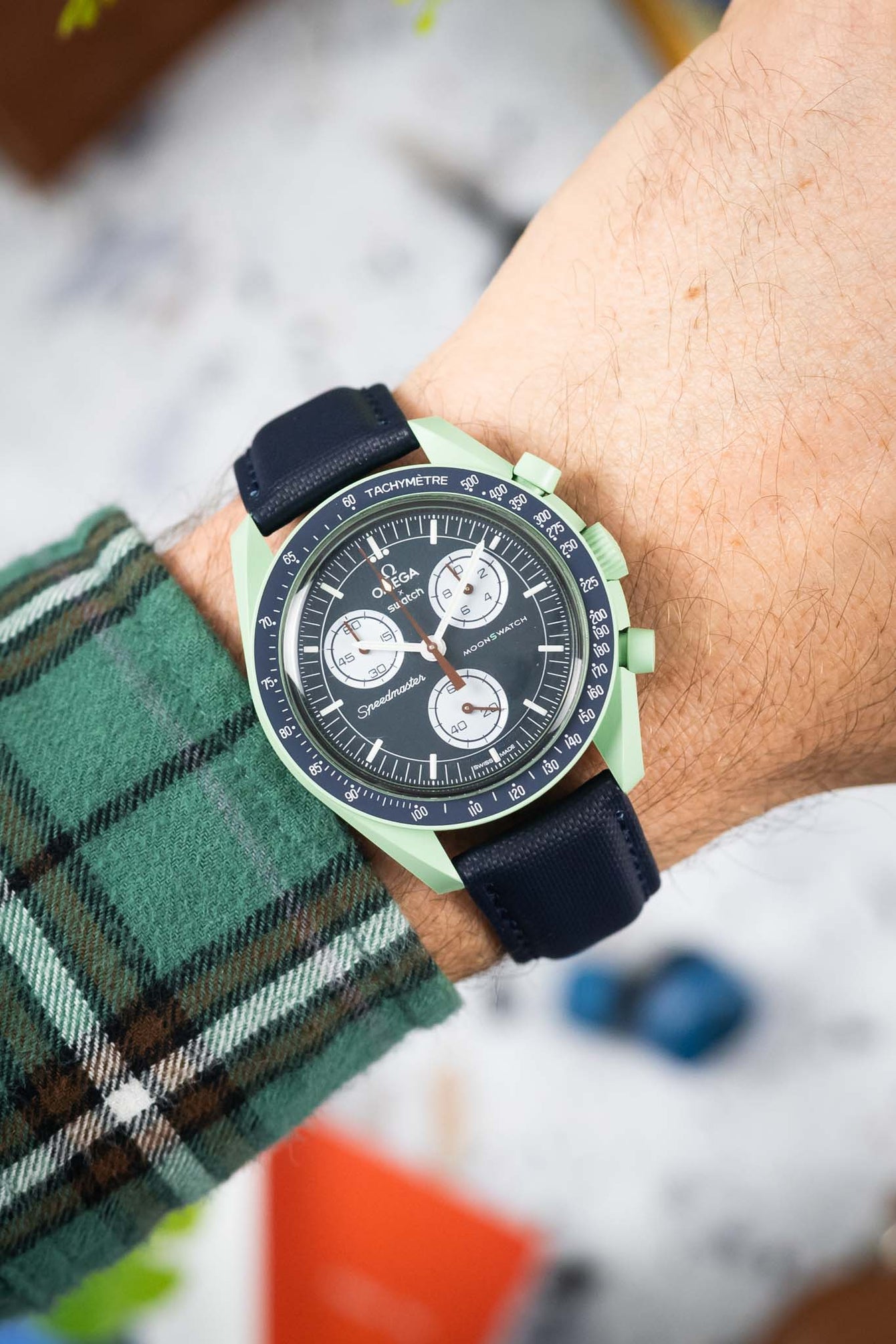 Close-up of a mint green Omega x Swatch Speedmaster-style watch with a black dial and white subdials, worn on a wrist with a dark textured strap. The tachymeter bezel and chronograph pushers are visible, while a green plaid sleeve frames the watch. The softly blurred background shows hints of orange and neutral tones, keeping the focus on the watch.