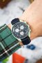 Close-up of a mint green Omega x Swatch Speedmaster-style watch with a black dial and white subdials, worn on a wrist with a dark textured strap. The tachymeter bezel and chronograph pushers are visible, while a green plaid sleeve frames the watch. The softly blurred background shows hints of orange and neutral tones, keeping the focus on the watch.