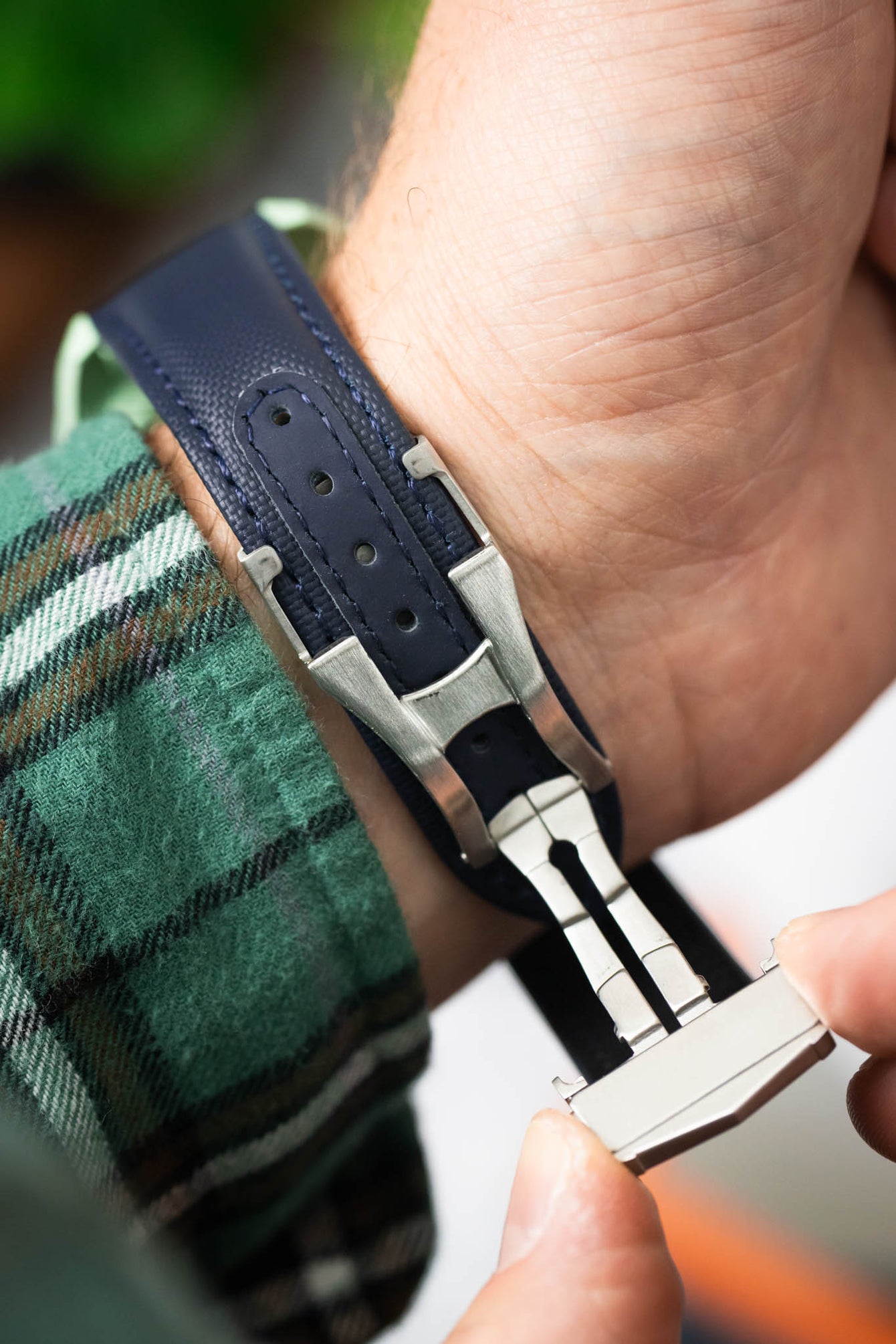 Close-up of a dark blue textured watch strap with a stainless steel deployant clasp being fastened on a wrist. The strap features visible stitching and adjustment holes, while the metal clasp is partially open, showing its folding mechanism as it is secured around the wrist.