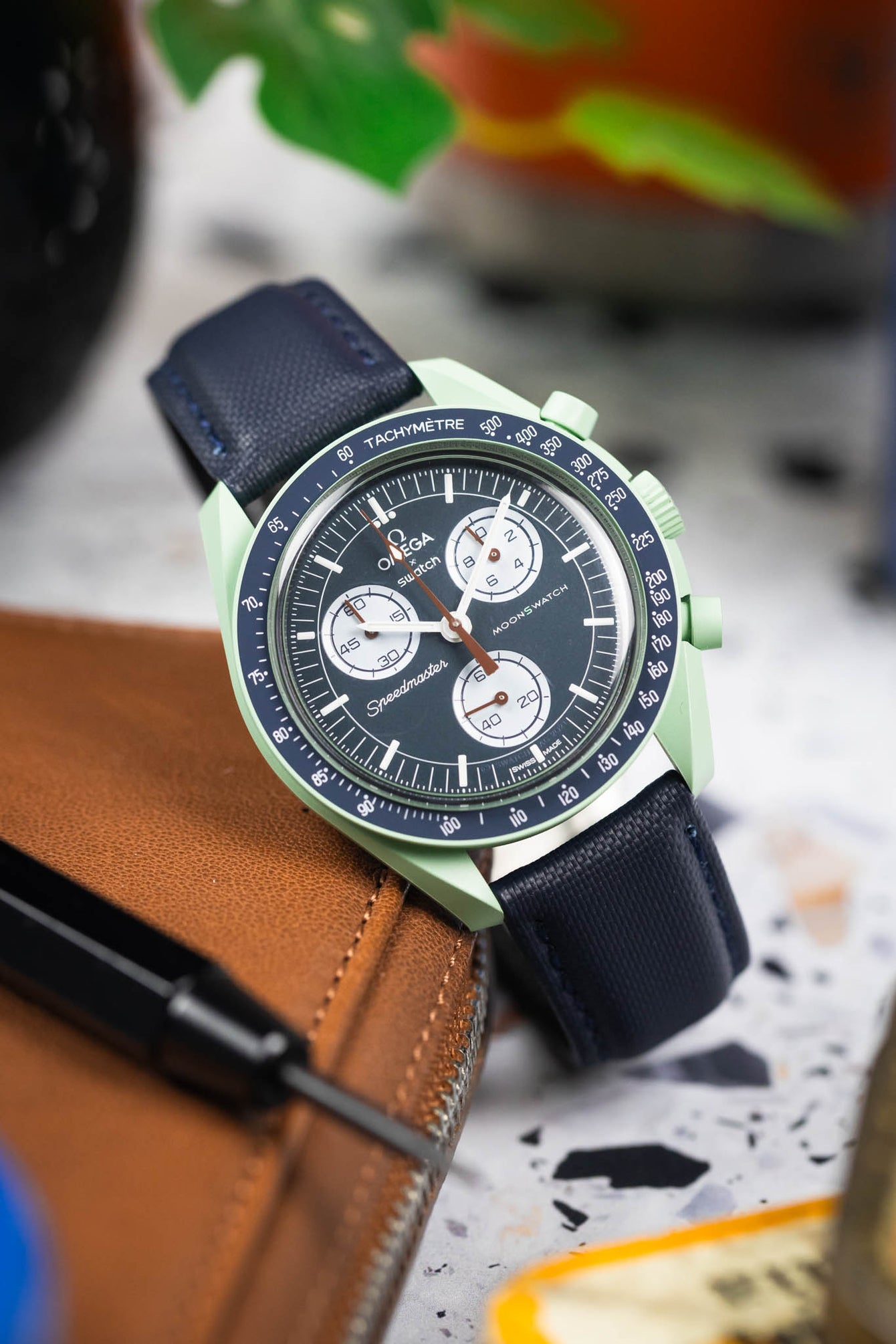 Close-up of a mint green Omega x Swatch Speedmaster-style watch with a black dial and white subdials, resting on a brown leather case. The watch is paired with a dark blue textured strap, while the softly blurred background features hints of greenery and warm tones that highlight the watch’s colors and details.