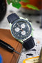 Close-up of a mint green Omega x Swatch Speedmaster-style watch with a black dial and white subdials, resting on a brown leather case. The watch is paired with a dark blue textured strap, while the softly blurred background features hints of greenery and warm tones that highlight the watch’s colors and details.