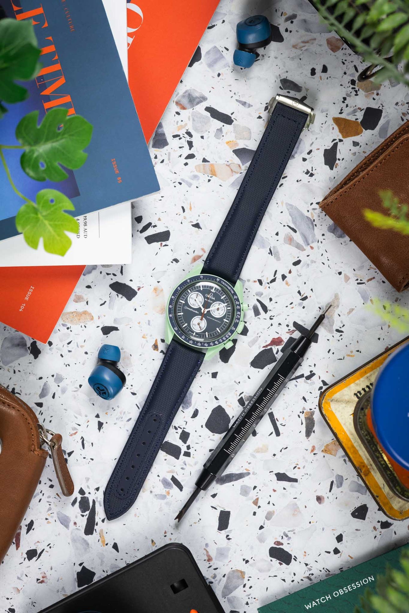 Flat lay of a green Omega x Swatch Speedmaster-style watch on a dark textured strap, placed on a speckled surface and surrounded by magazines, a tool, earbuds, a leather pouch, and plants.