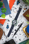 Flat lay of a green Omega x Swatch Speedmaster-style watch on a dark textured strap, placed on a speckled surface and surrounded by magazines, a tool, earbuds, a leather pouch, and plants.