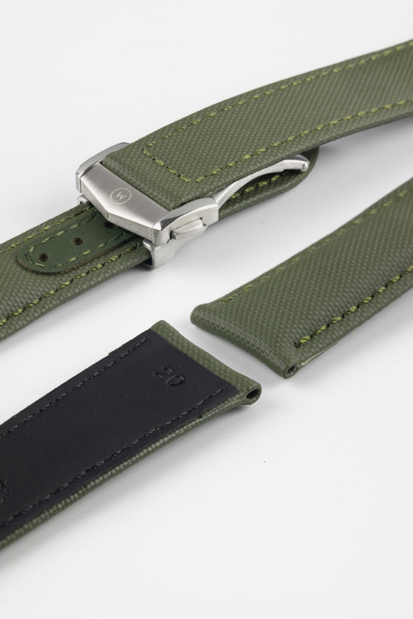 Angled close-up of an olive green textured watch strap with matching green stitching, featuring a brushed silver deployant clasp with an engraved logo, alongside separate strap sections, all arranged on a light grey background.