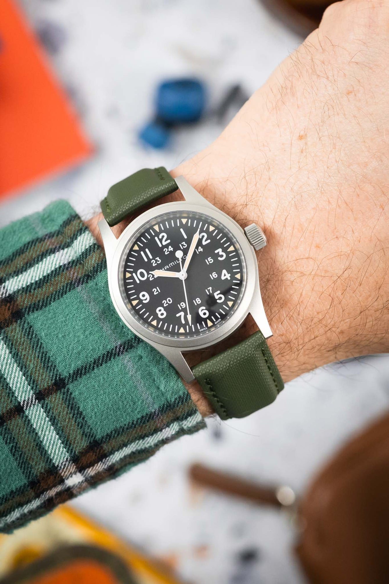 Close-up of a Hamilton-style field watch with a black dial and full Arabic numerals, worn on a wrist with a green textured strap. The dial features 24-hour markings and luminous hands, while a green plaid sleeve and softly blurred workshop-style background frame the watch.